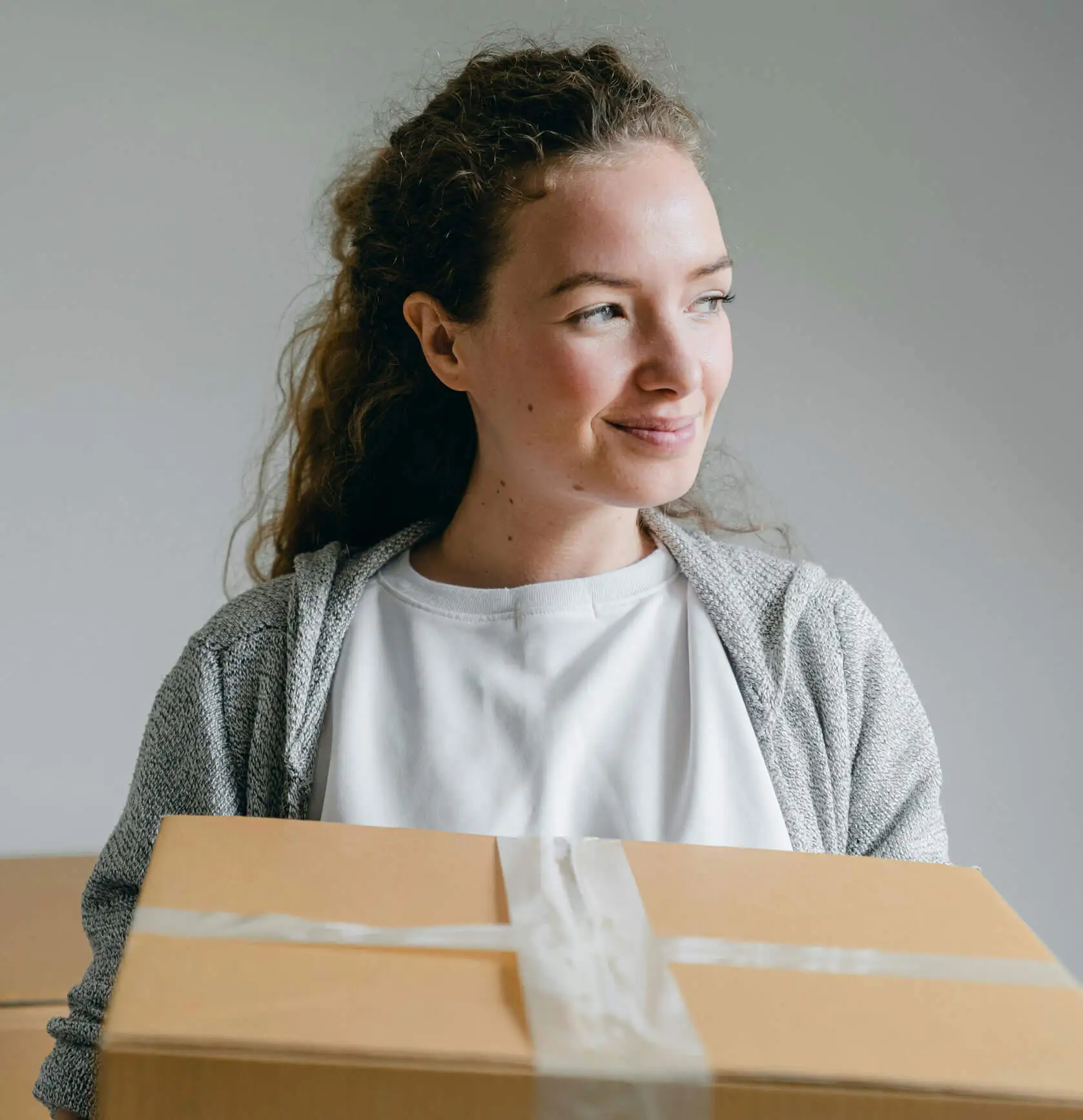A woman with short, curly brown hair stands in front of a grey wall, holding a cardboard box in front of her. She is wearing a blue sweater and jeans.